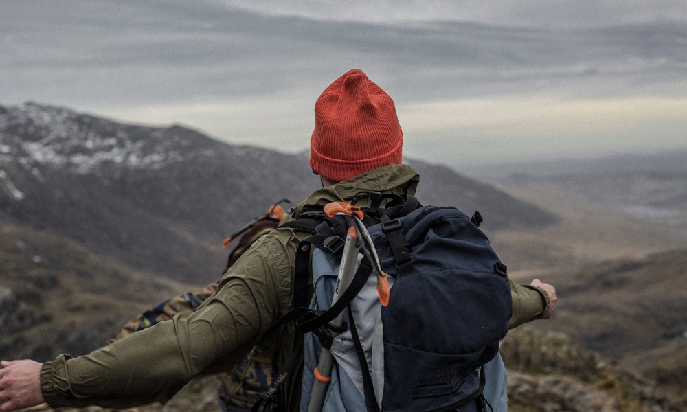 A man in beanie and backpack standing atop of a mountain with his arms stretched out.