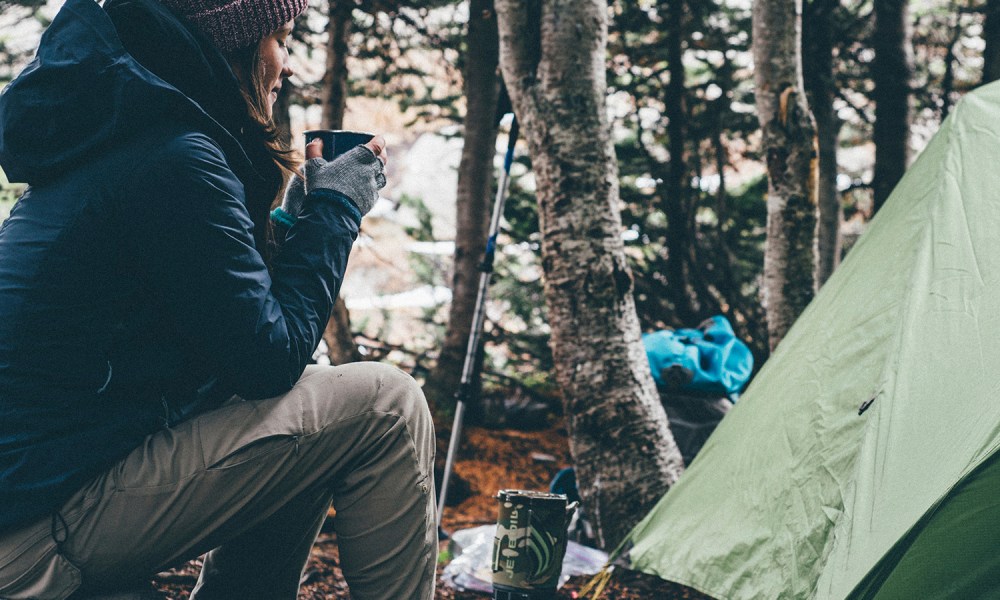 A woman sitting and drinking a warm beverage by a green tent.