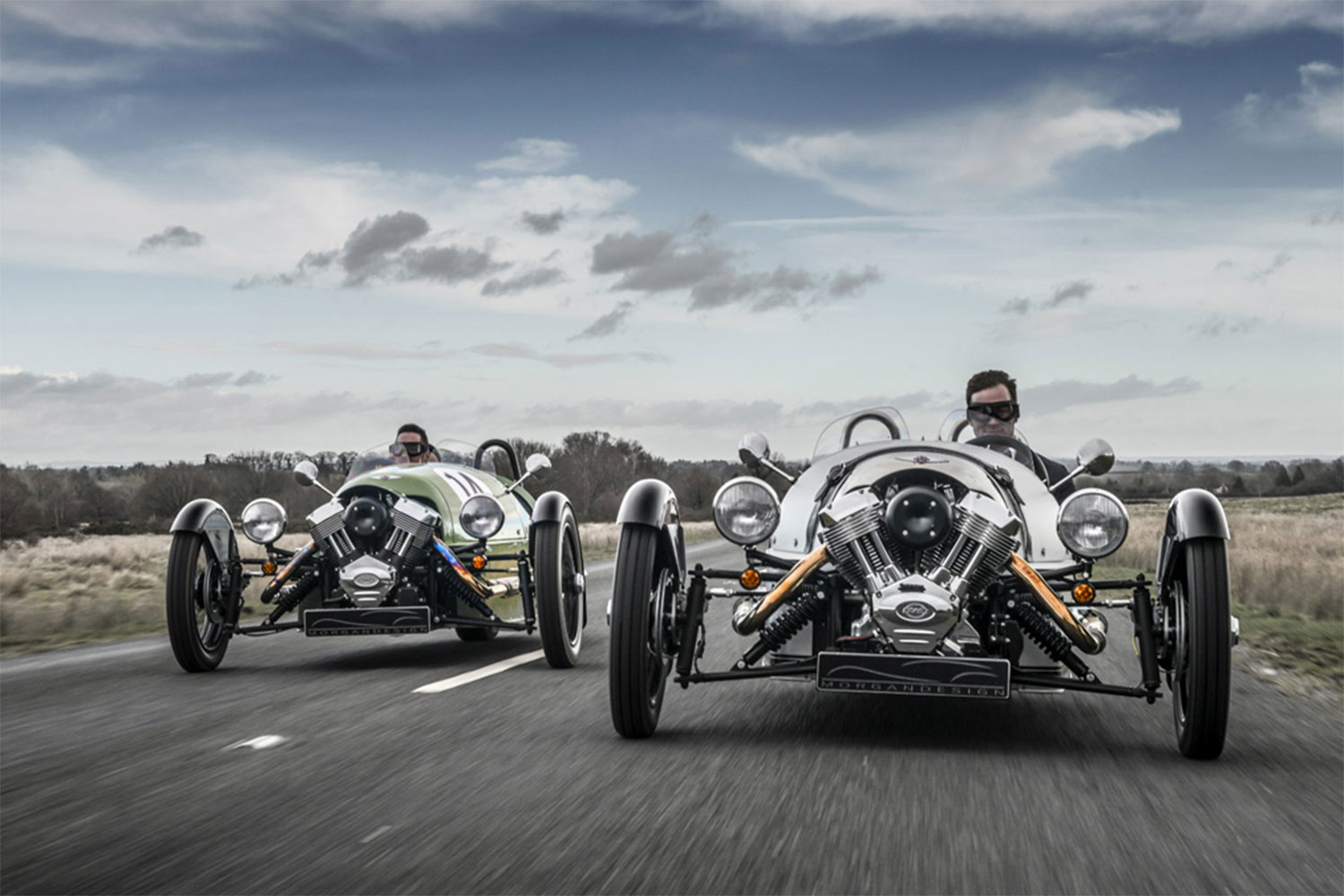 Two Silver Morgan 3-Wheeler being driven in a wide road.