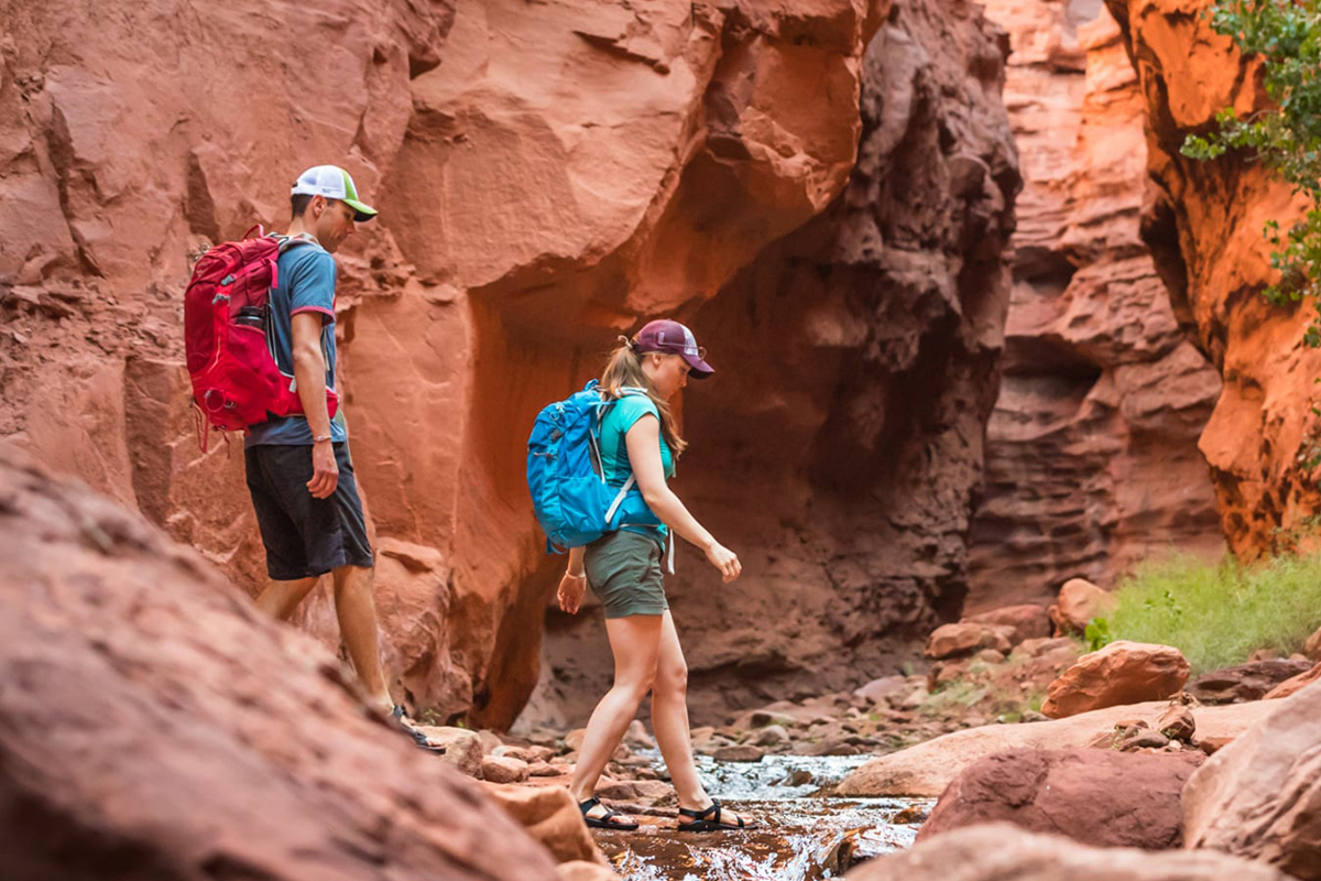 Two hikers in summer clothing cross a small creek in the red bedrock