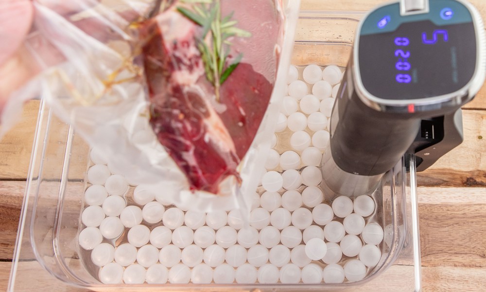 A hand holding a vacuum-sealed bag containing meat on a tray with a sous vide machine.