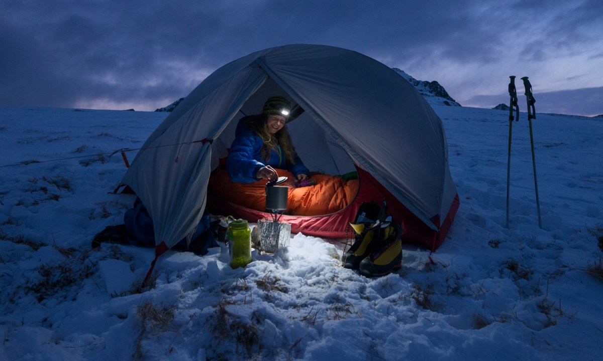 Woman with a headlamp camping in a winter tent surrounded by snow.