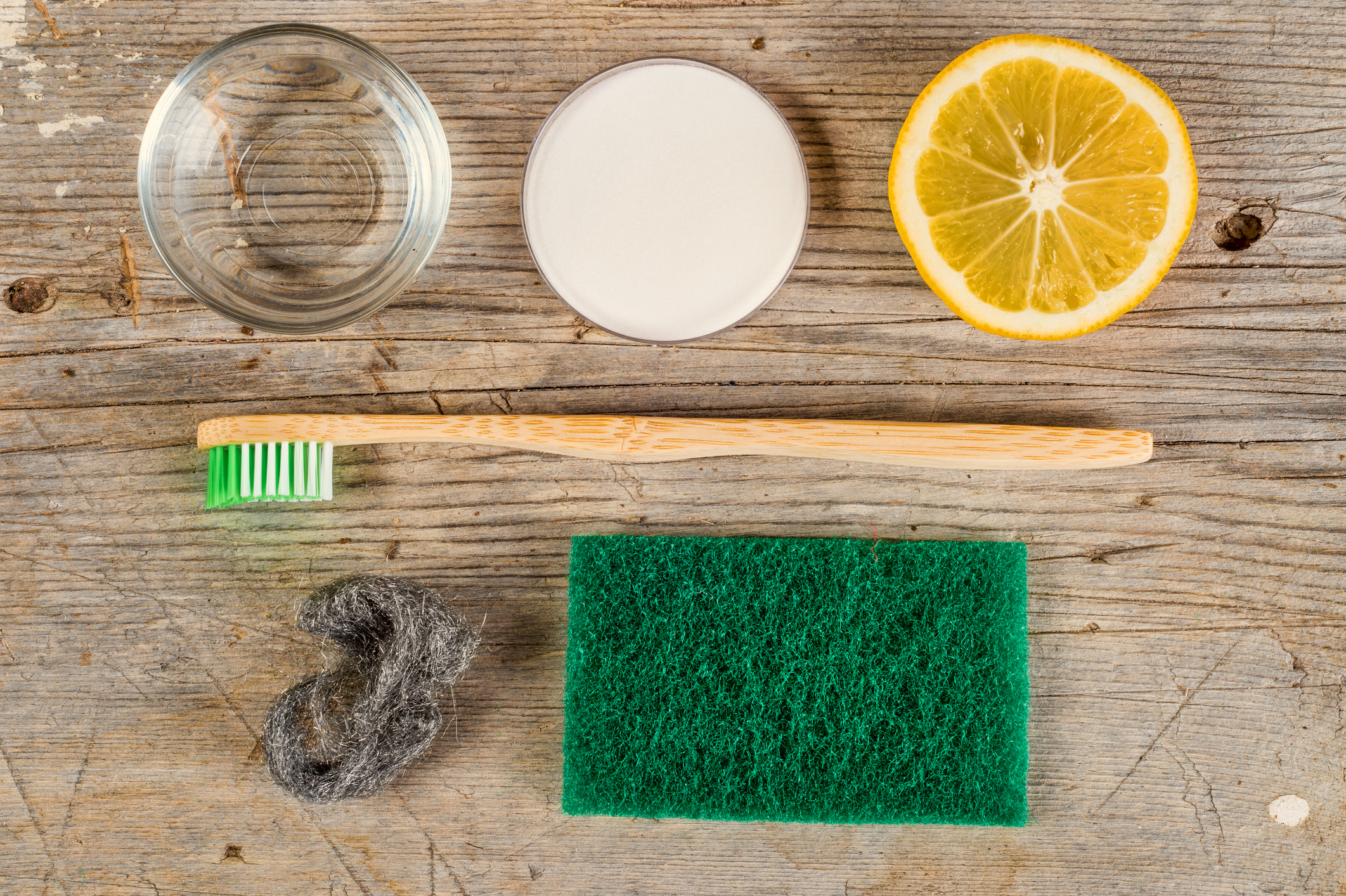 An overhead shot of a wooden toothbrush, sponge, lemon, soap, baking soda, steel wool, and a jar of water on wooden surface.