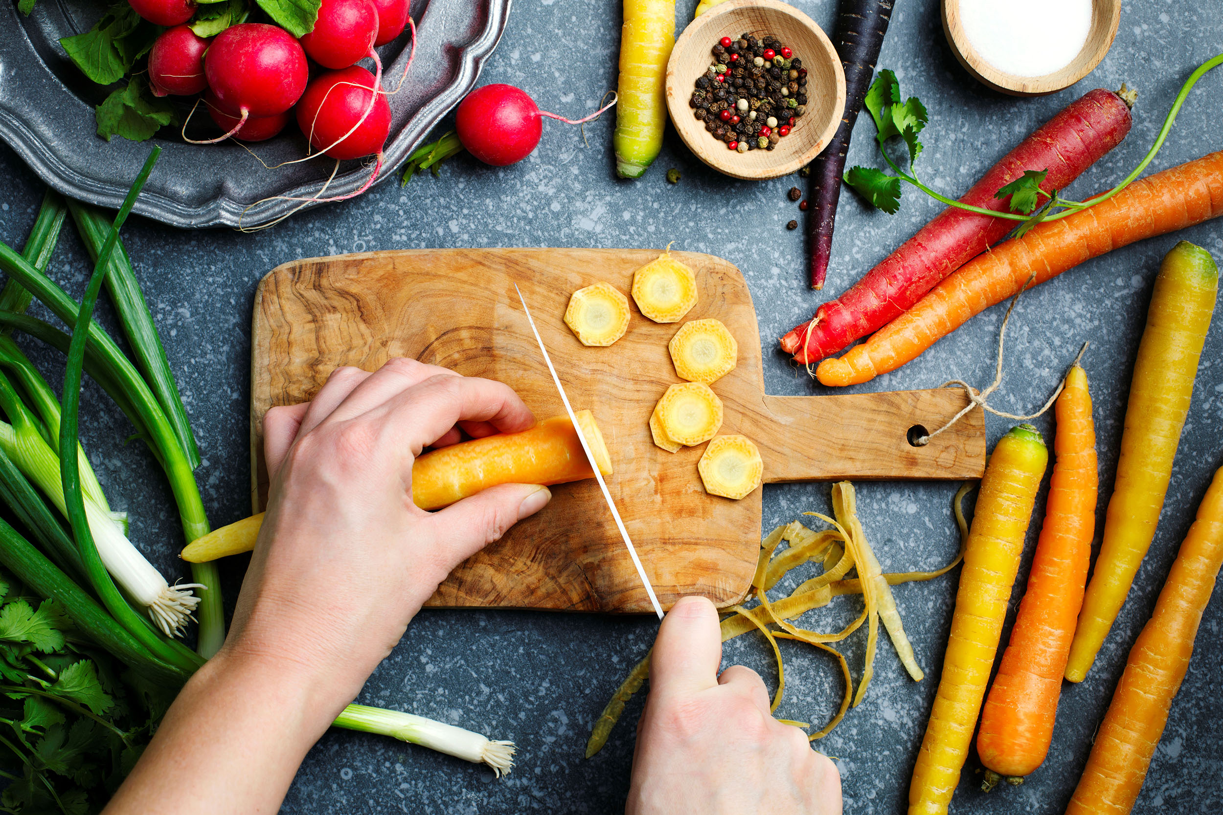 Cutting carrots on wooden board and fresh spring vegetables for vegetarian cooking on the table, top view.