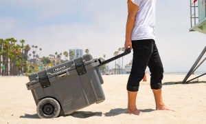 A man hauling a gray wheeled Pelican cooler on a beach.