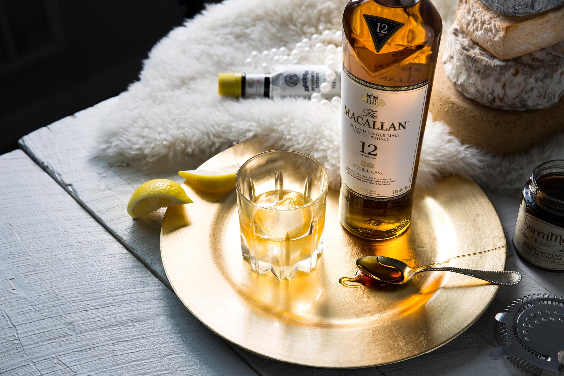 A bottle of The Macallan, a cup of Honeypot cocktail, lemon slices, and a spoonful of syrup on top of a golden plate on a wooden surface.