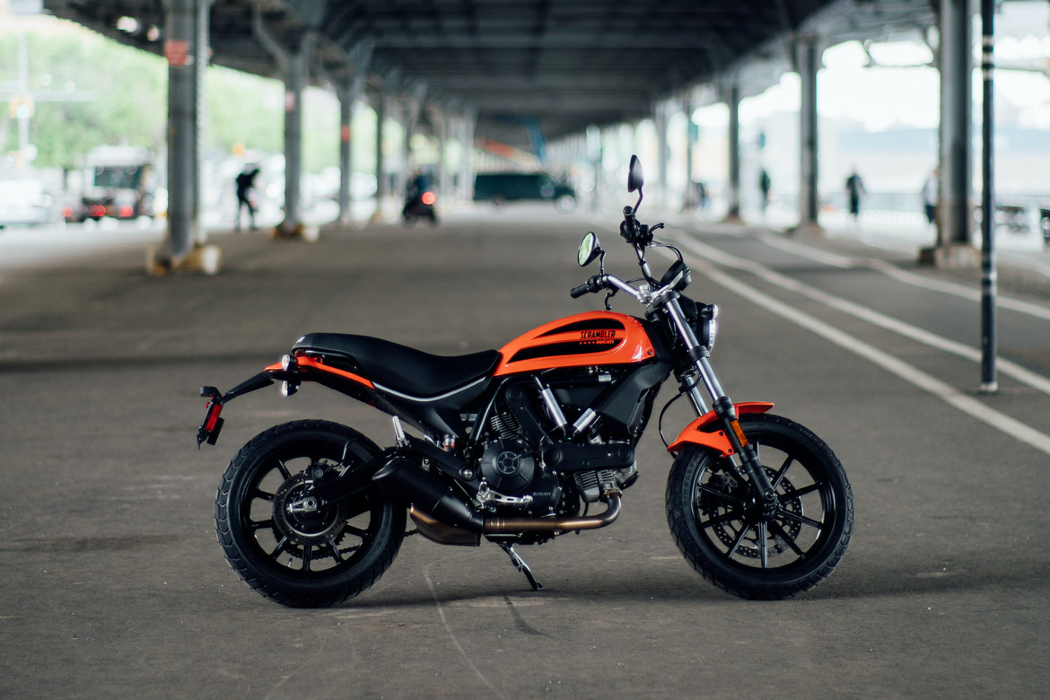 Ducati Scrambler Sixty2 underneath a bridge with pedestrians and cars in the back.