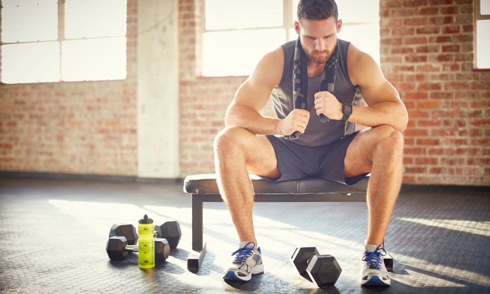 A tired man with towel sitting on a bench in a gym.