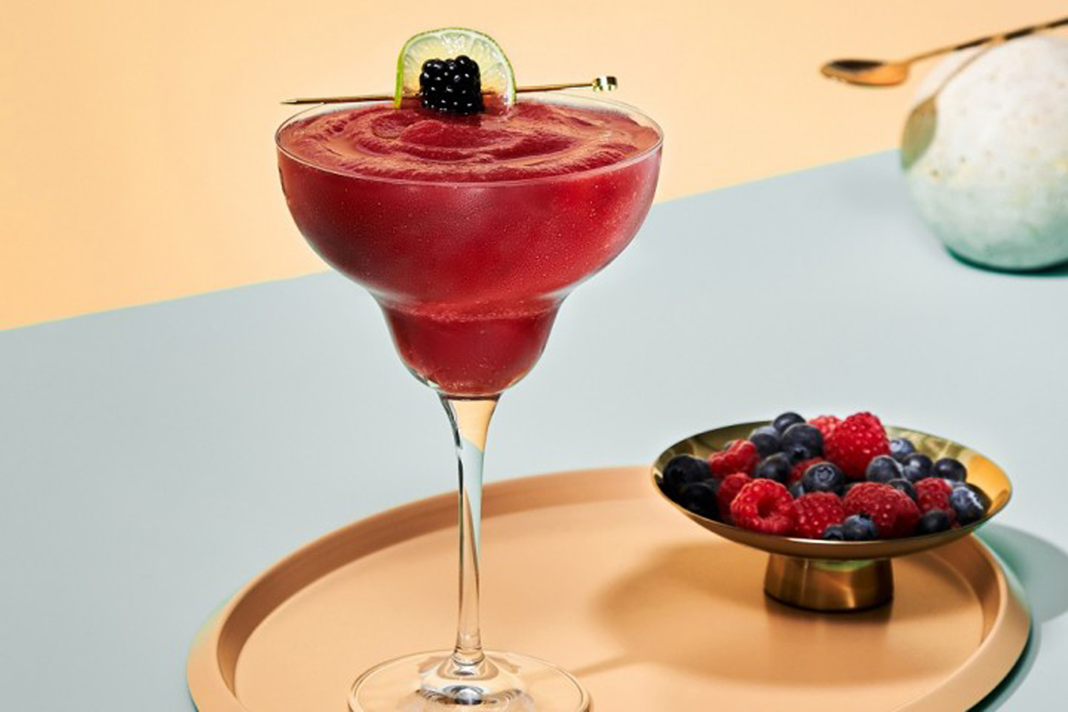 A glass of Frozen Berry Margarita beside a bowl of berries on a circular tray.