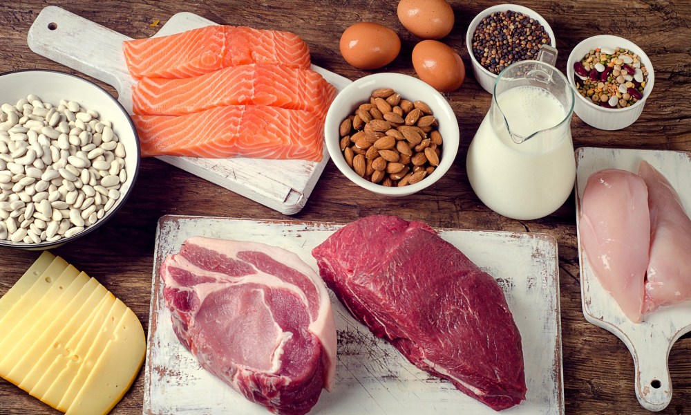 Fish fillets, chicken meat, and red meat on top of distressed white cutting boards along with nuts, cheese, dairy, and eggs