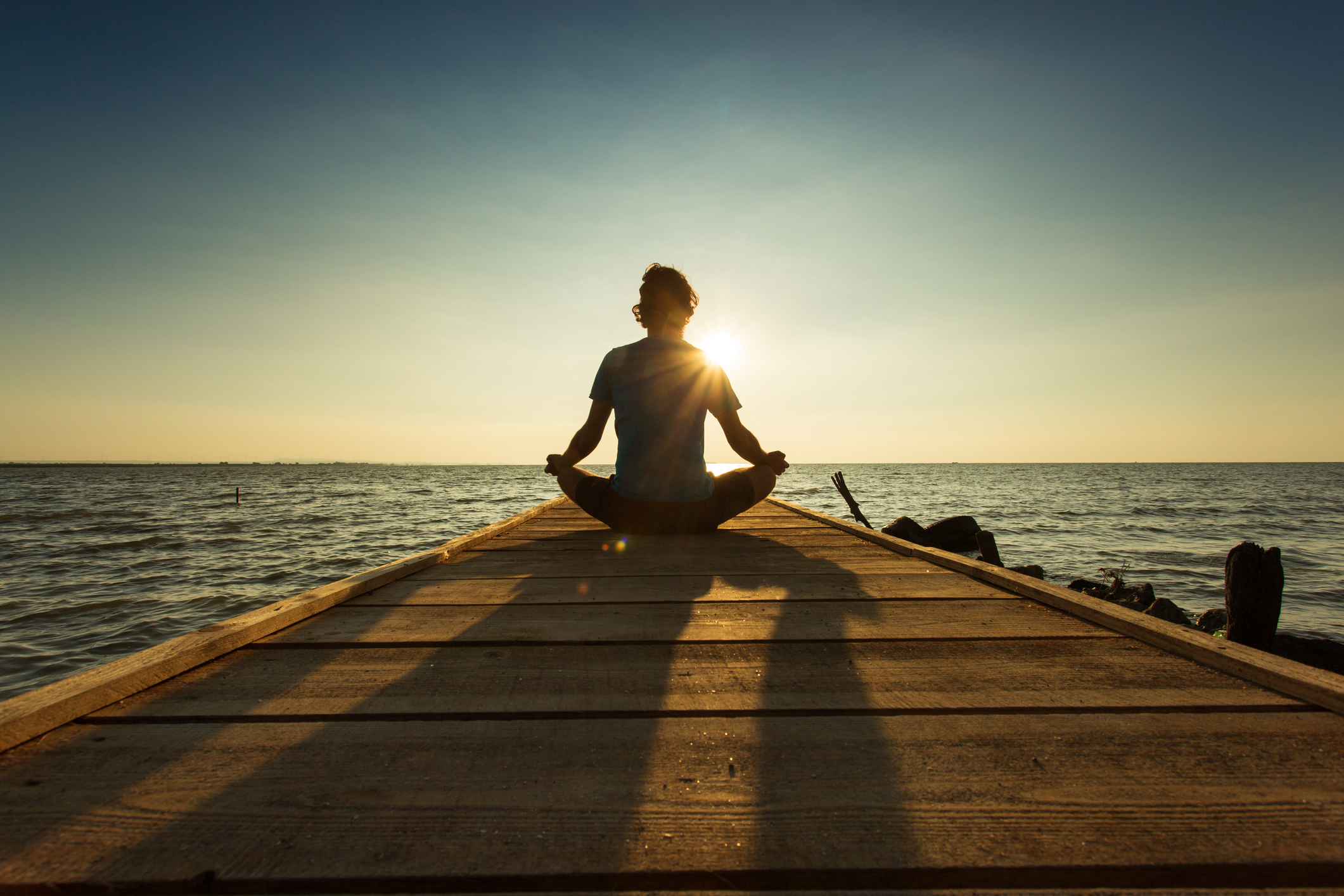 Young man sitting and meditating on a pontoon by a lake at the sunrise.