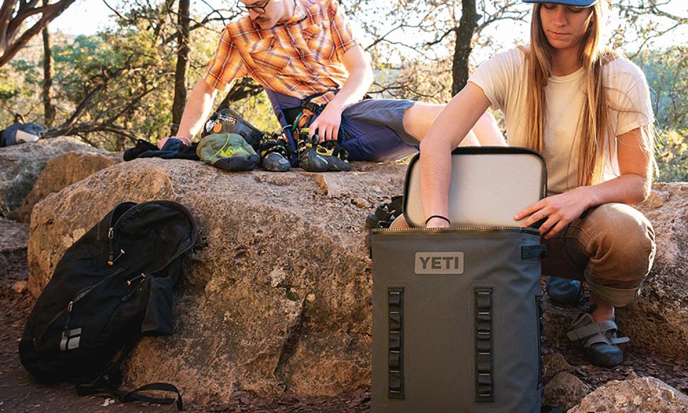 A man on a boulder with outdoor gear and an opened backpack while a woman is fishing out a drink from a Yeti cooler.