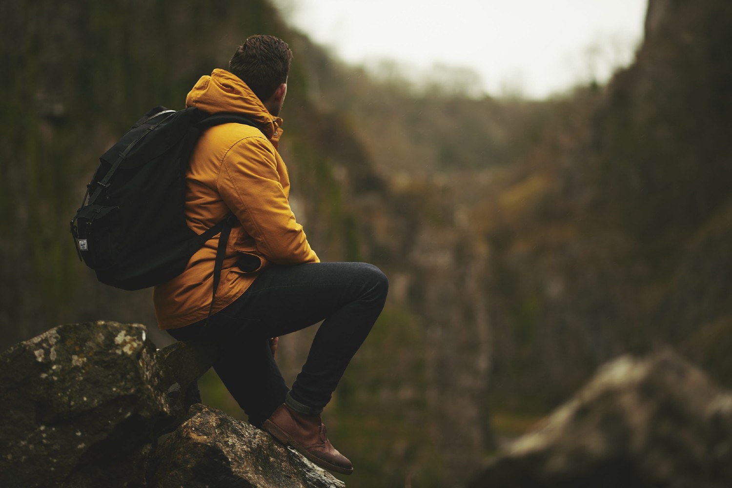 A hiker in a yellow waterproof sits on a rock and looks uphill on an overcast day