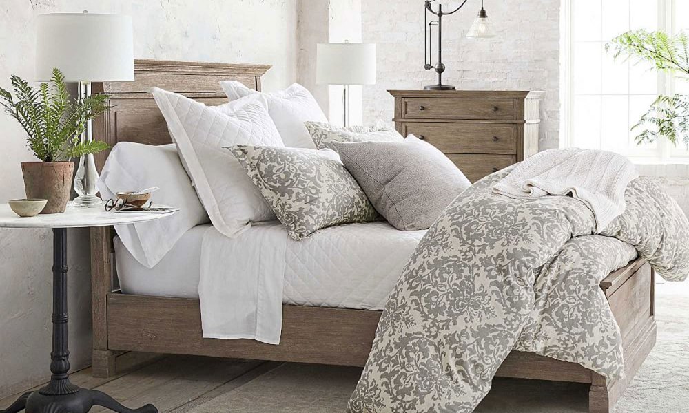 A wooden bed with pillows and a comforter near a bedside table with a potted plant and decor in a bedroom.