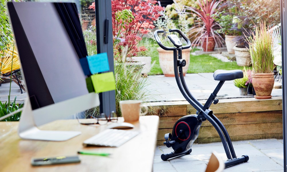 A folding exercise bike beside a table with desktop computer in a home office.