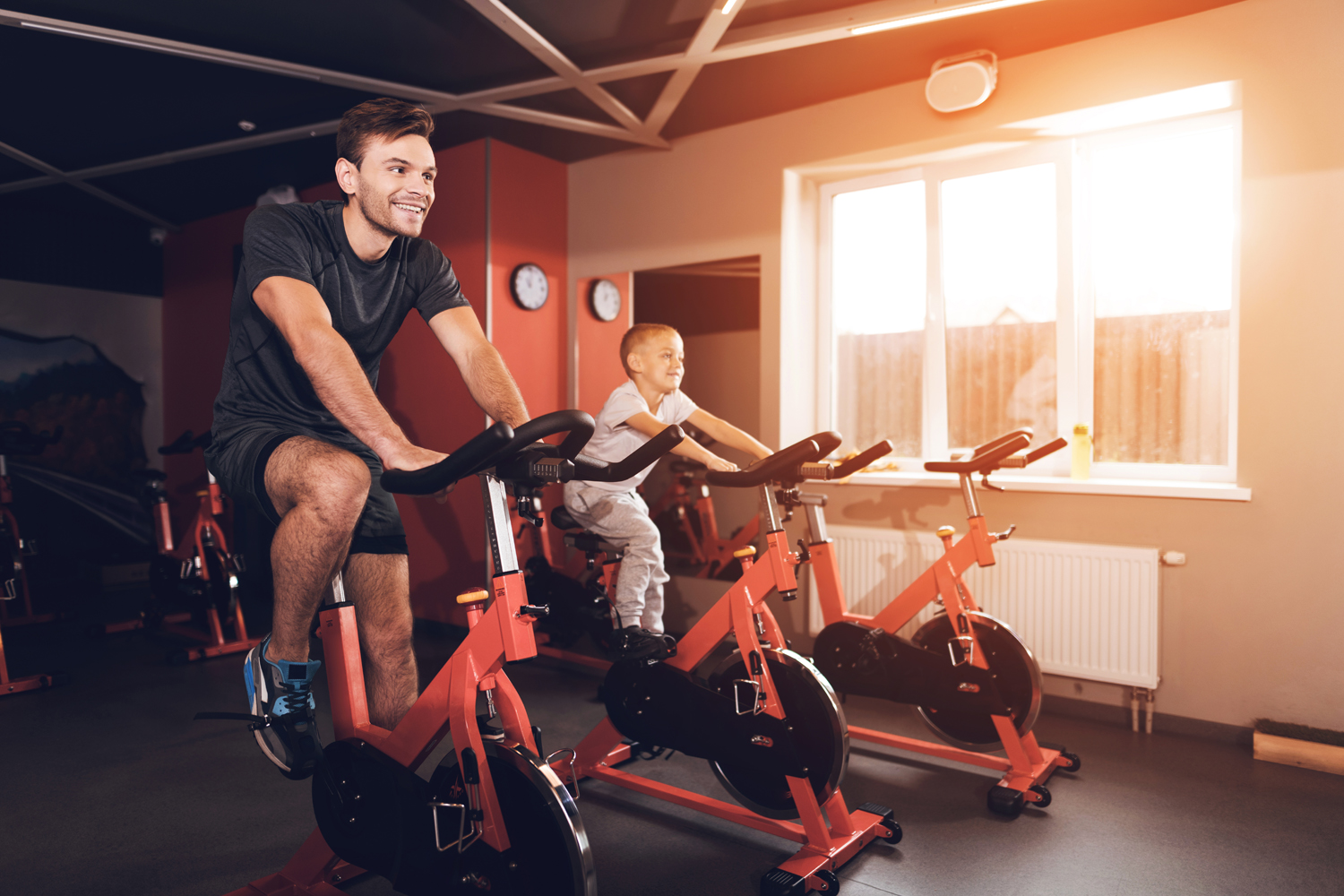 A father and his son using exercise bikes in a gym on a sunny day.
