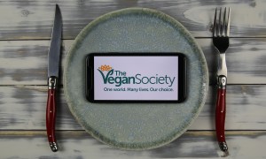 The logo of The Vegan Society displayed on a phone on a plate, with a knife and fork beside it on a table.