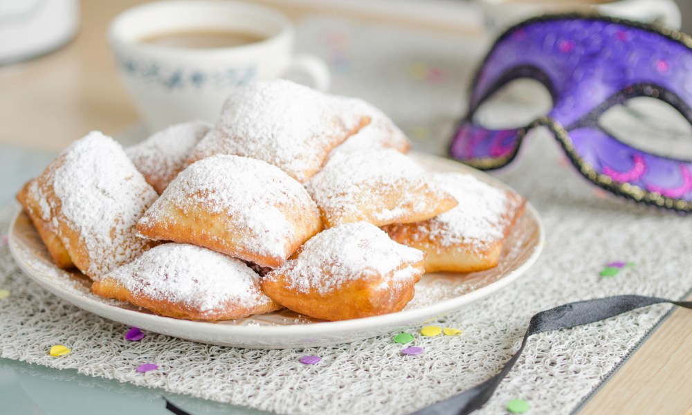 Beignets topped with sugar on a plate with a purple mask, a cup of hot drink, and confetti.