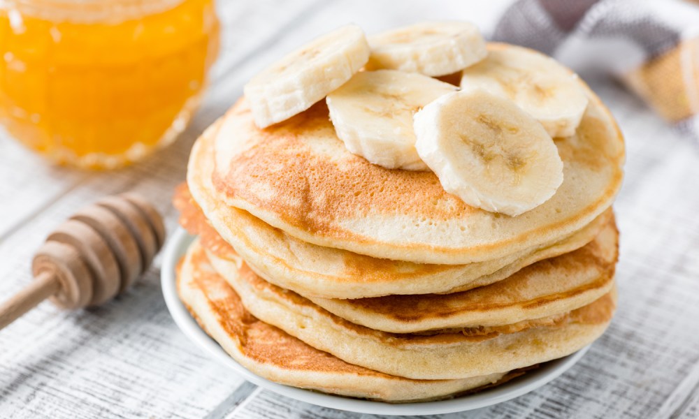 Banana slices on a plate of pancakes beside a cup of honey on a table.