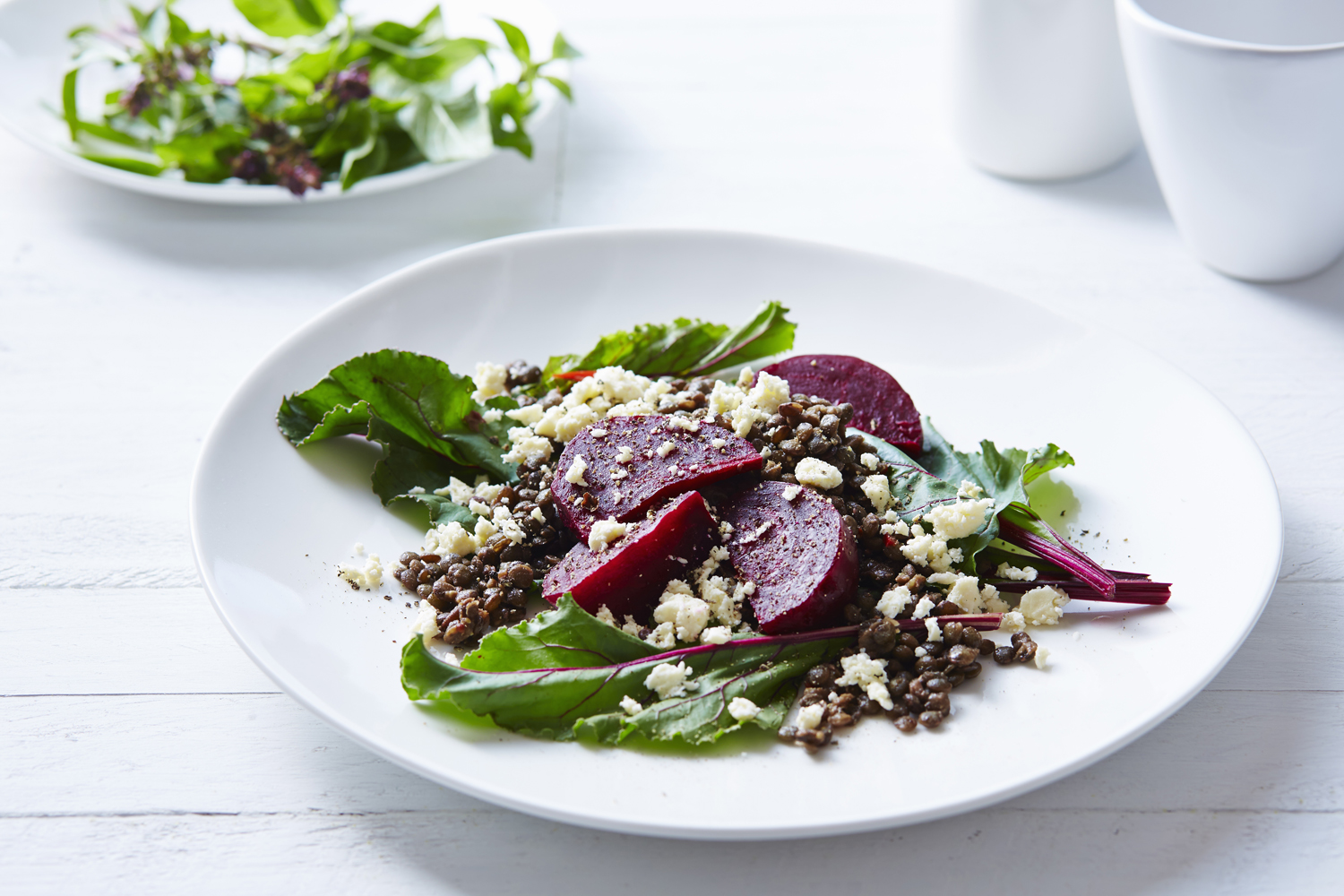 Two plates of beetroot and feta salad beside two cups on white table.