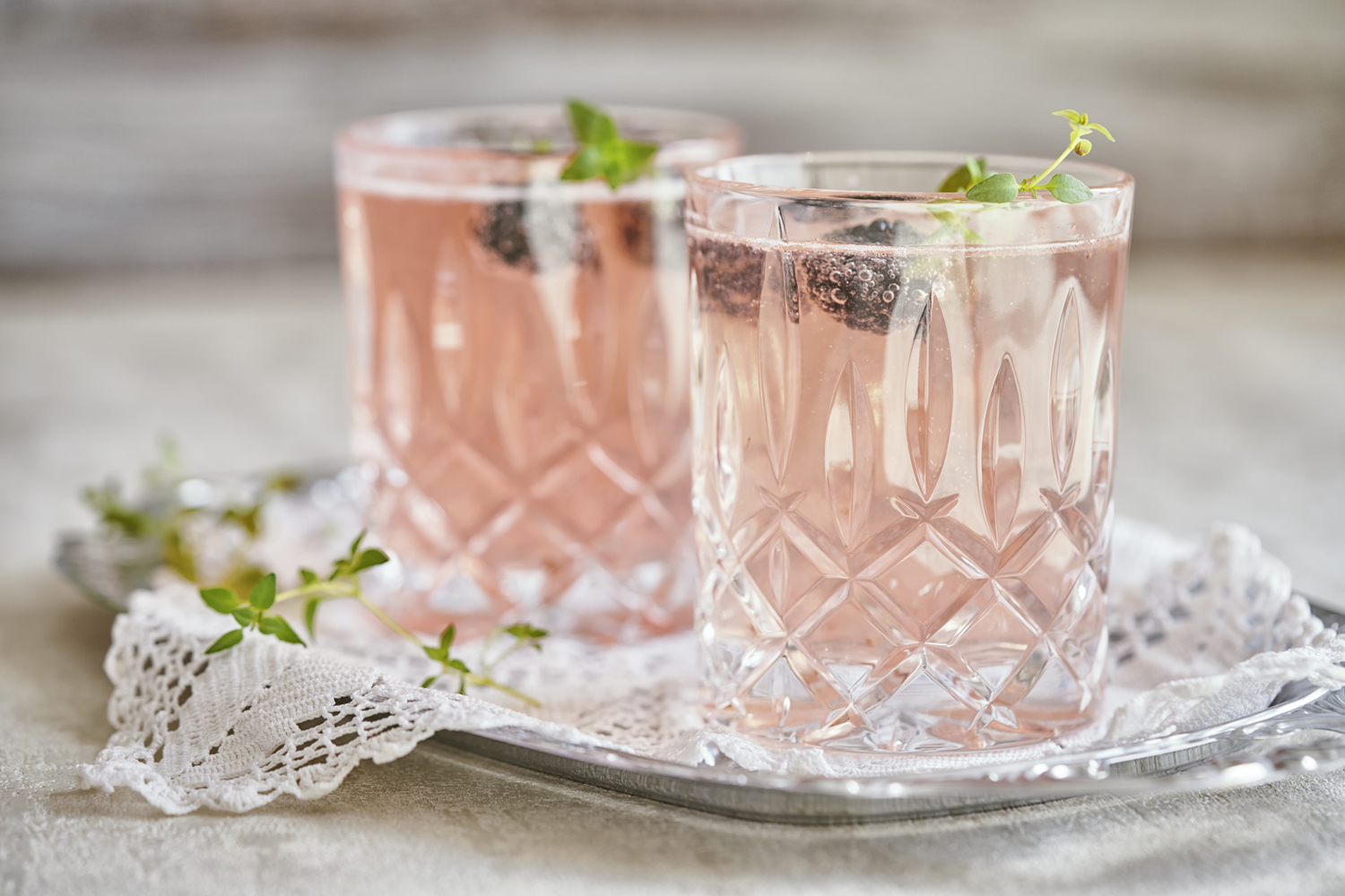 Two glasses of Rose cocktails with blueberry garnish on crocheted cloth on a metal tray