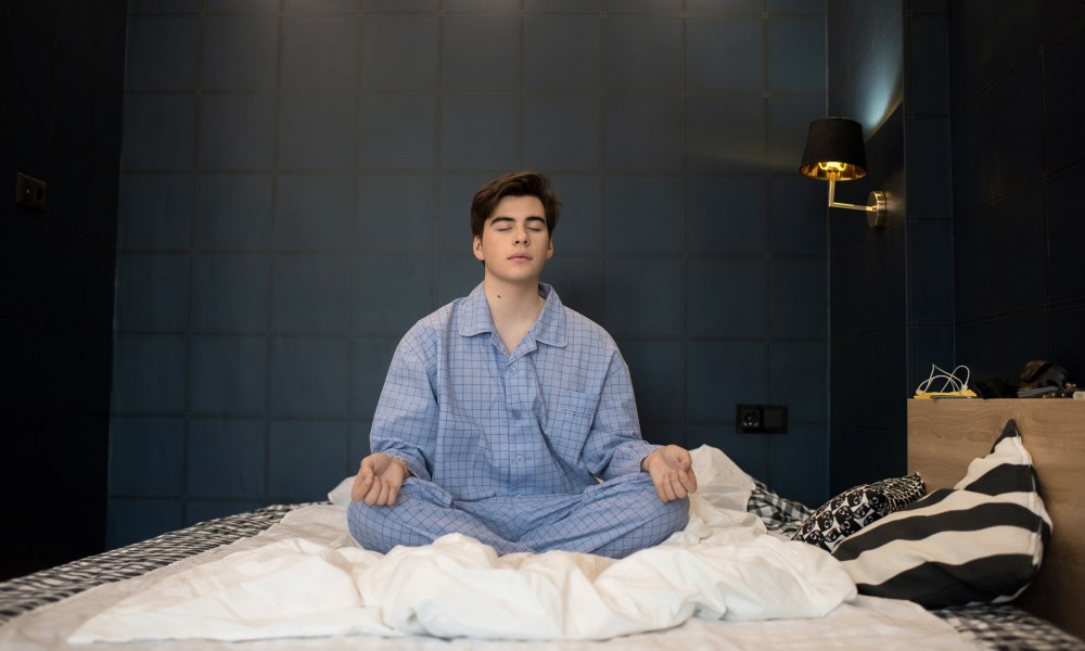 Portrait of teenage boy wearing pajamas meditating with eyes closed while sitting on bed in lotus position trying to sleep at night