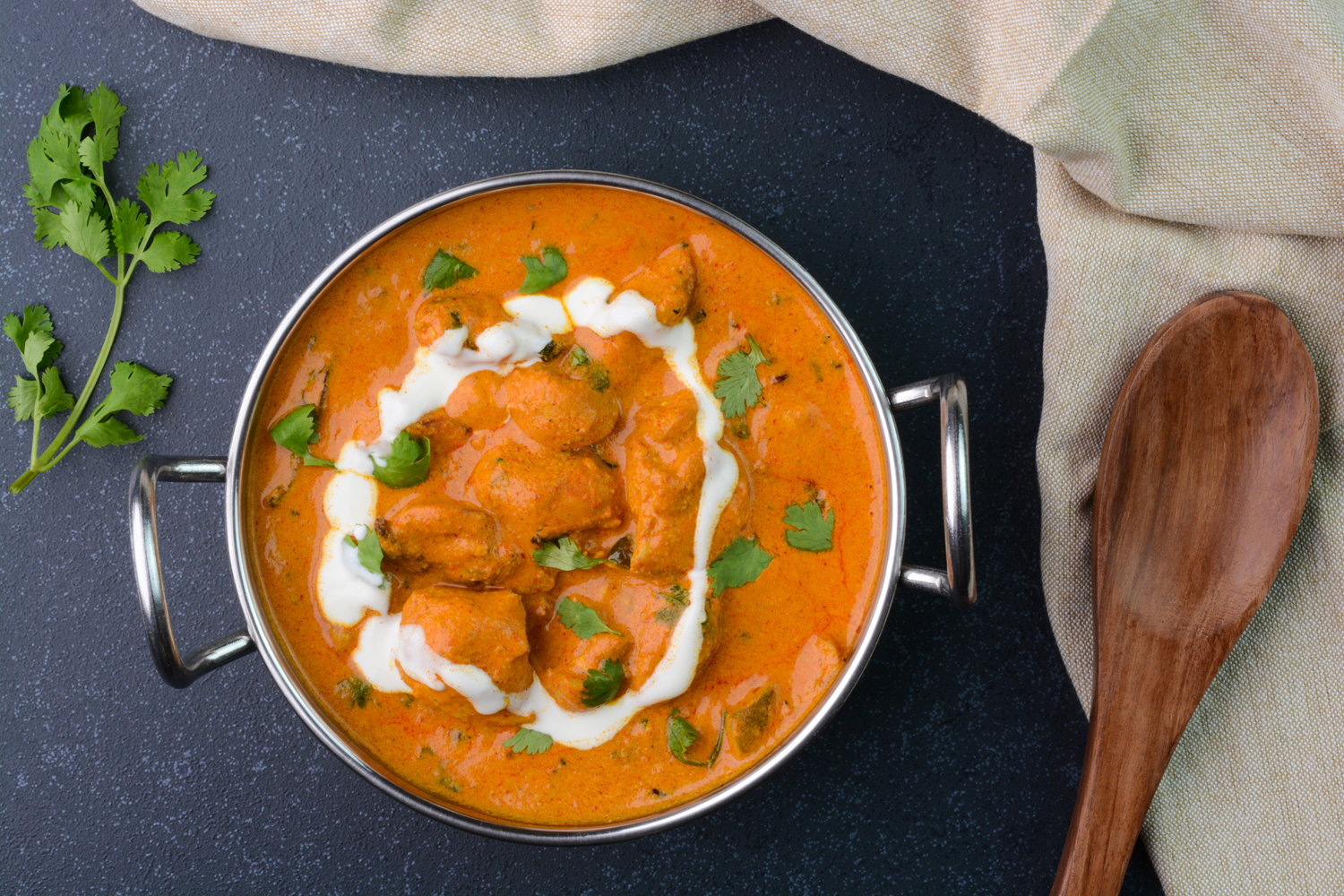 An overhead shot of butter chicken in a pot beside herbs and a wooden spoon.