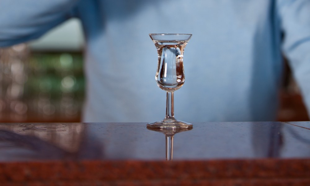 A glass of Genever on bar counter with a midsection of a man behind it.