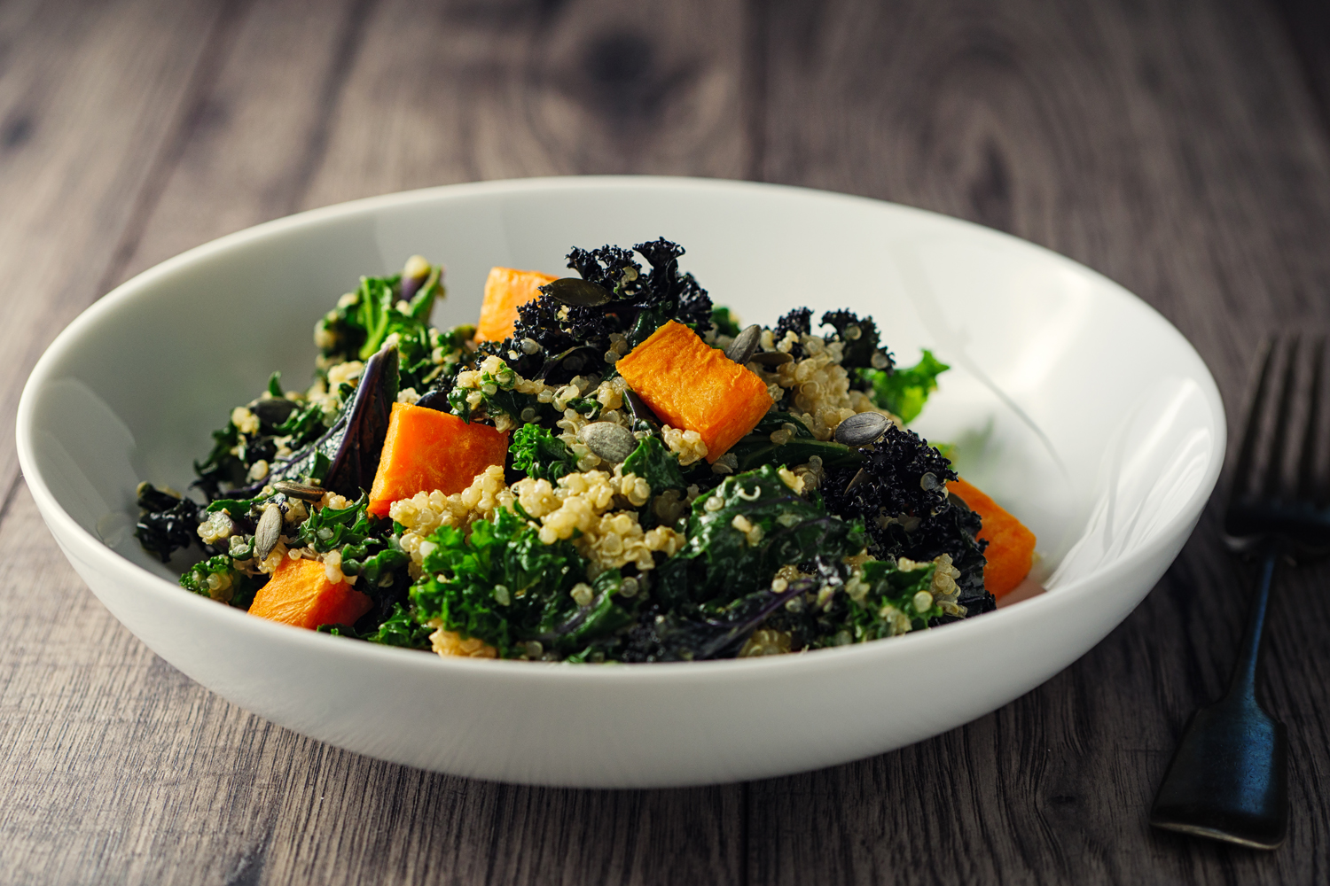 A bowl of kale salad with roasted sweet potatoes and pumpkin seeds beside a fork on a wooden table.