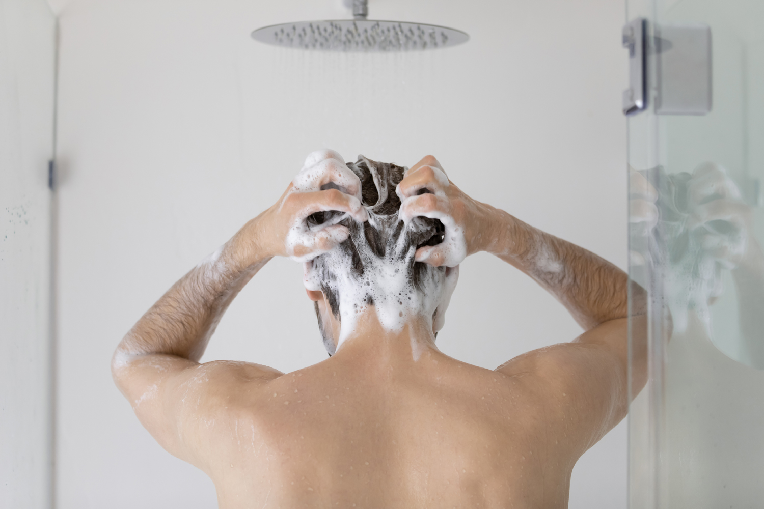 A rear view of a man lathering shampoo on his hair in the shower.