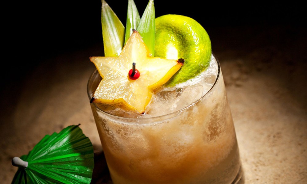 A Mai Tai cocktail topped with fruits and leaves beside a cocktail umbrella