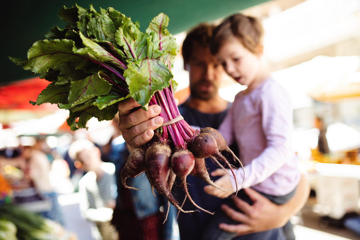 A father holding a bunch of beets while carrying his son in the market.