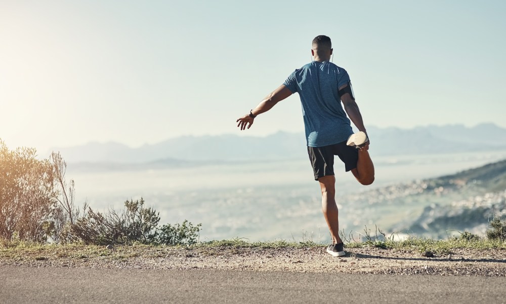 A man balancing on one leg doing a quad stretch, on a road with a view of the mountain