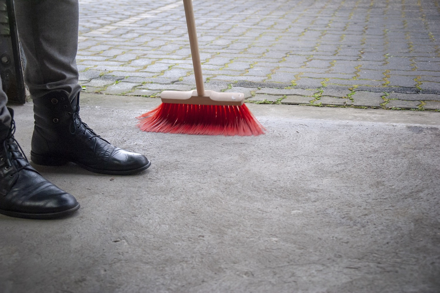 A person sweeping dirt on a concrete floor with a red brush.