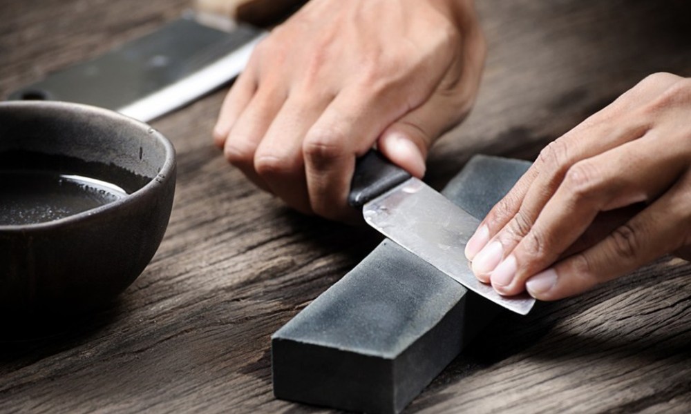 Hands sharpening a knife using a whetstone beside a bowl of water on a wooden table