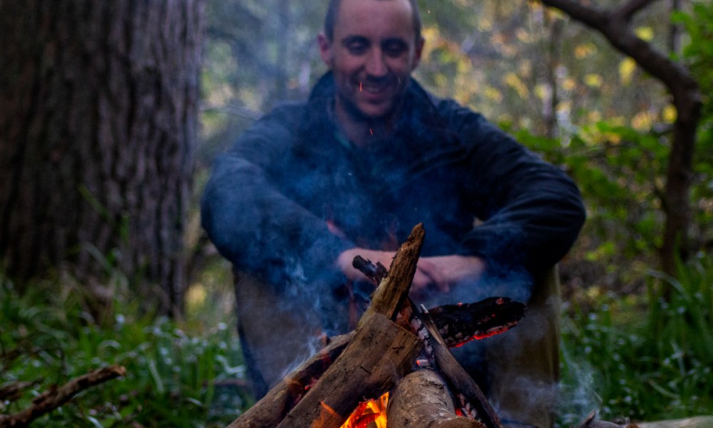 A man sits on the far side of the campfire while smoke rises in front of him.