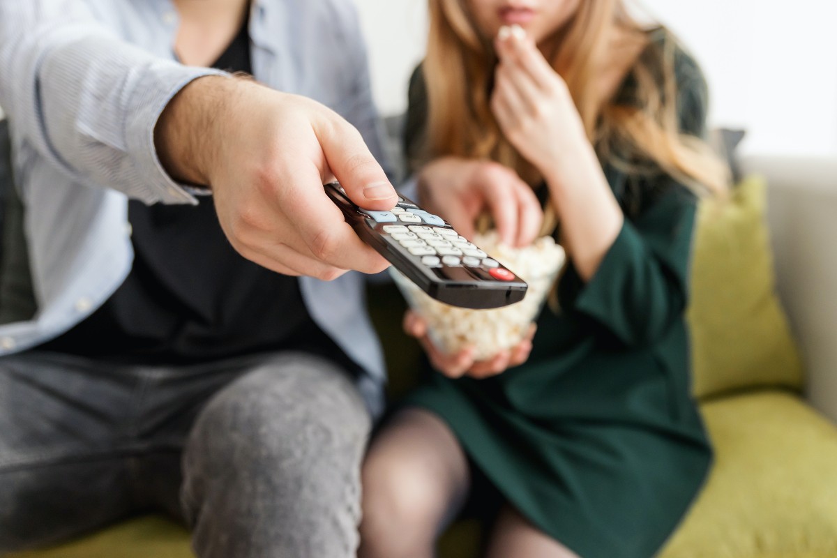 two people watching TV on the couch. one person has a remote. another is eating popcorn.