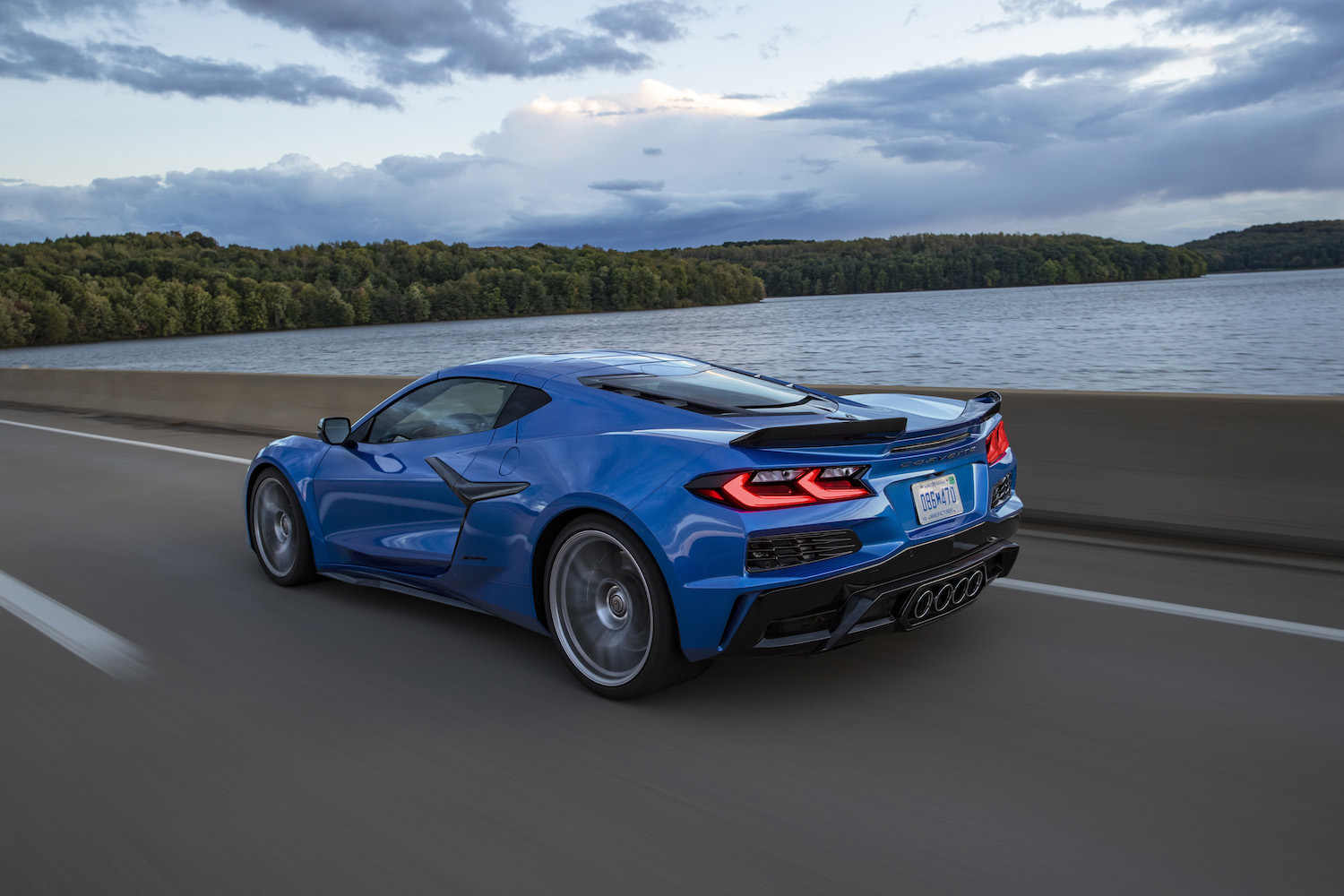 2023 Chevrolet Corvette Z06 rear end angle from driver's side driving on the road in front of a body of water with clouds in the back.