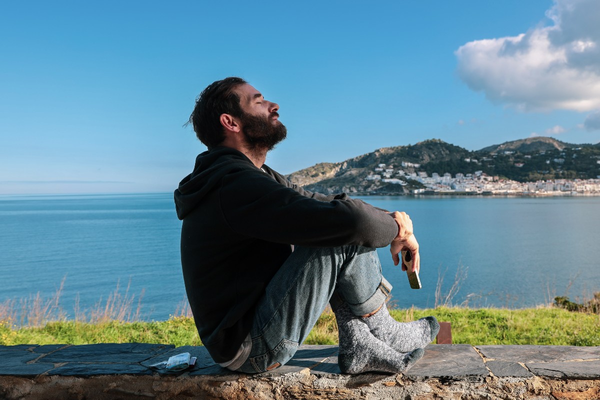 Sad man sitting on the ground near a body of water with his eyes closed.