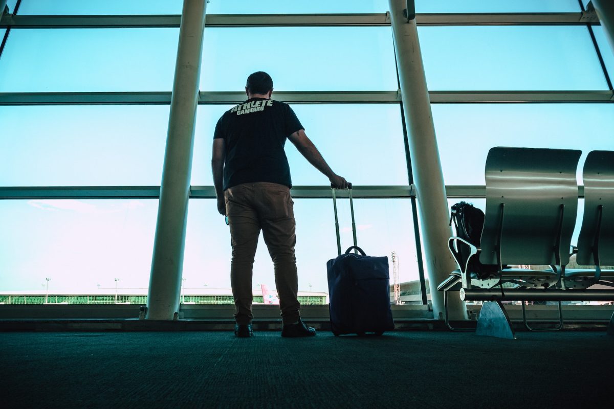 A man holding his luggage while looking out the window at the airport.