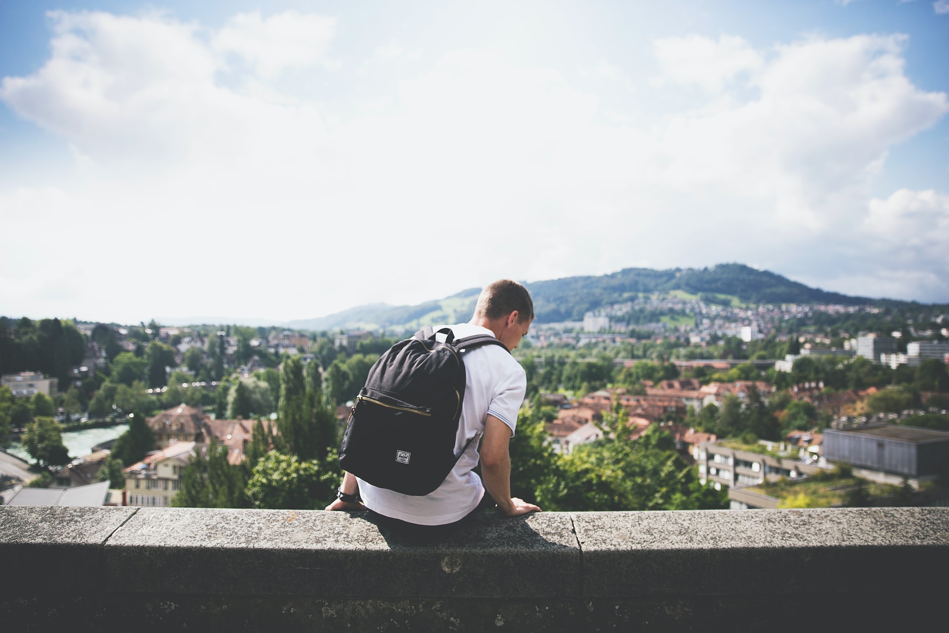 Man with a backpack sitting on a concrete wall overlooking a mountain and urban landscape