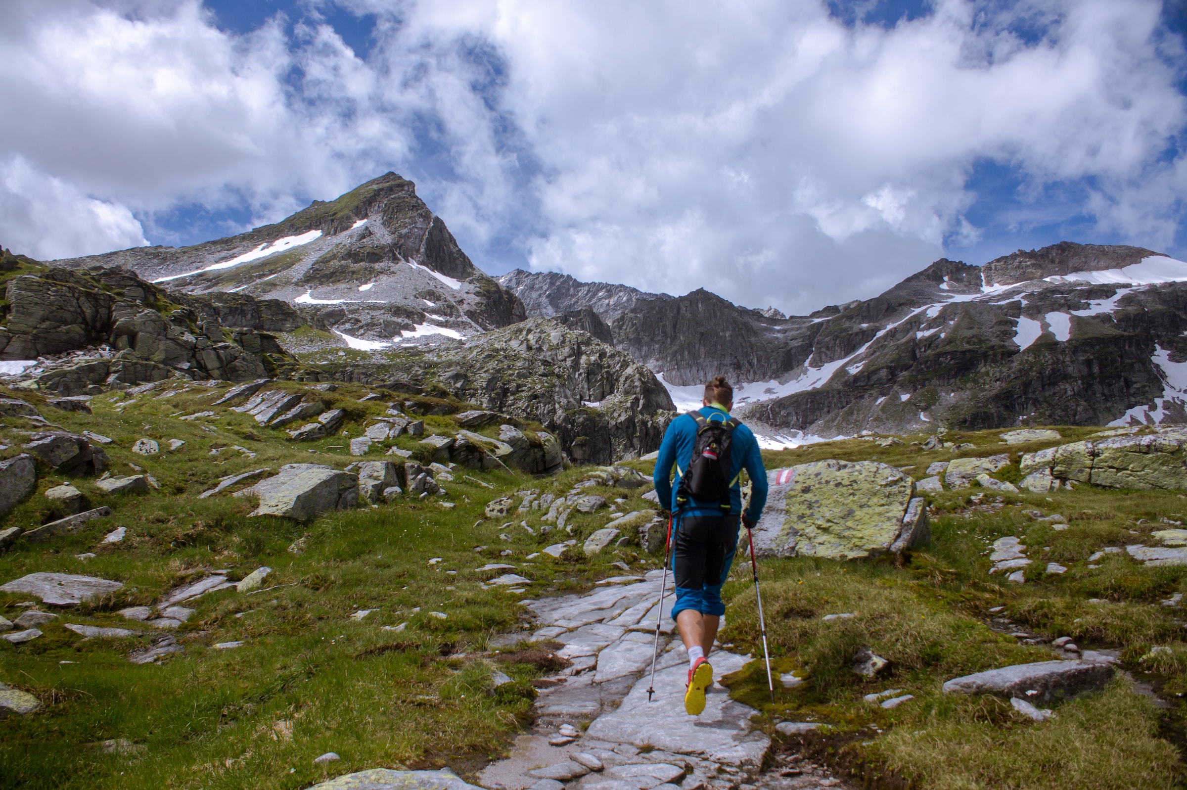 A man hikes up a trail towards a set of snow-topped mountains.