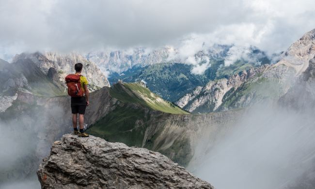 A man with a red backpack stands on a mountaintop looking over more mountains.