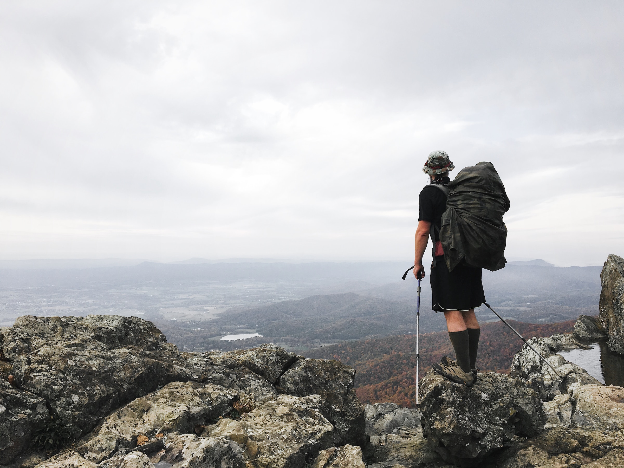 A man stands high up on a trail with hiking poles and a backpack.