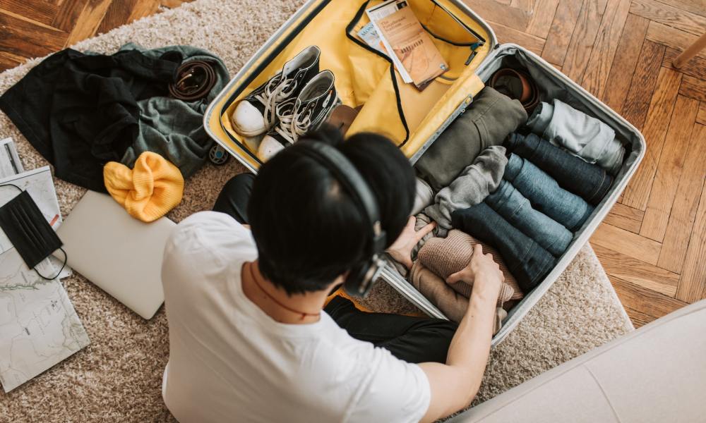 Top view of a man wearing headphones while packing a suitcase with clothing