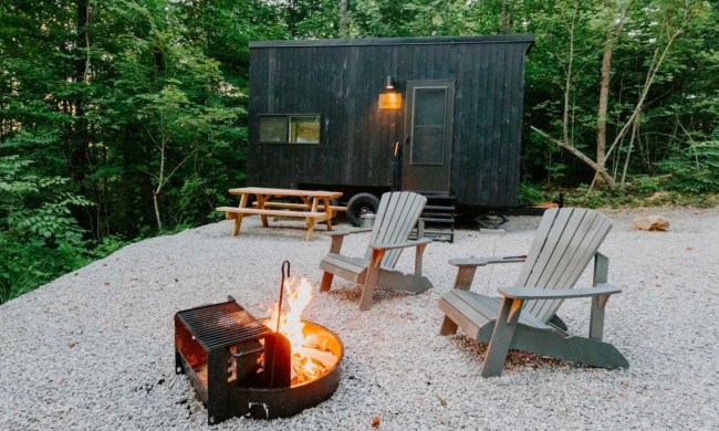 A small, black wood-paneled tiny house in the woods with a firepit and chairs in the foreground
