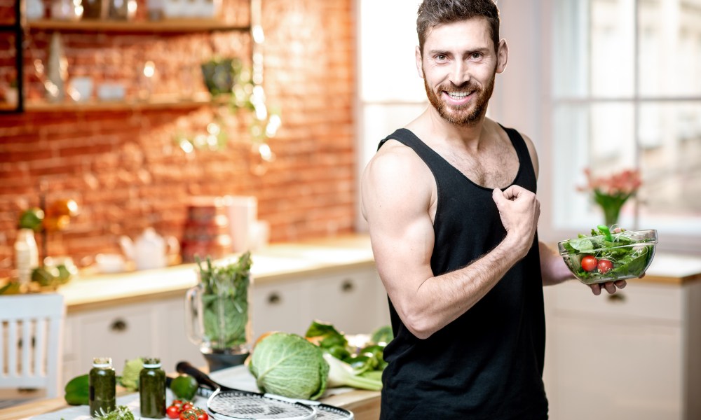 Portrait of a handsome sports man showing muscles, eating healthy vegetarian salad on the kitchen at home