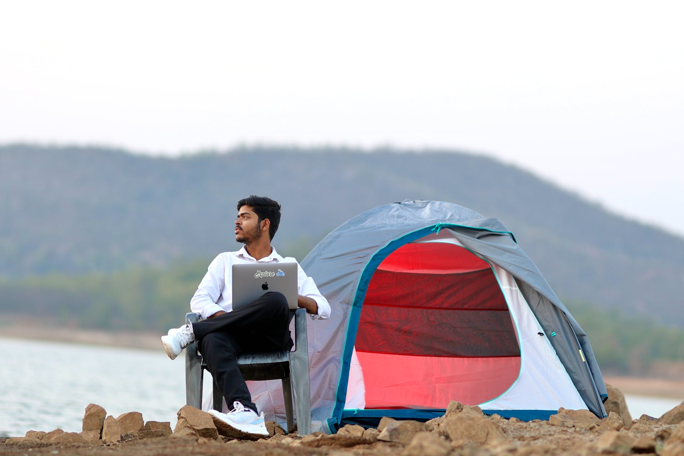 A man sitting while holding a laptop at his campsite off a body of water on a cloudy day