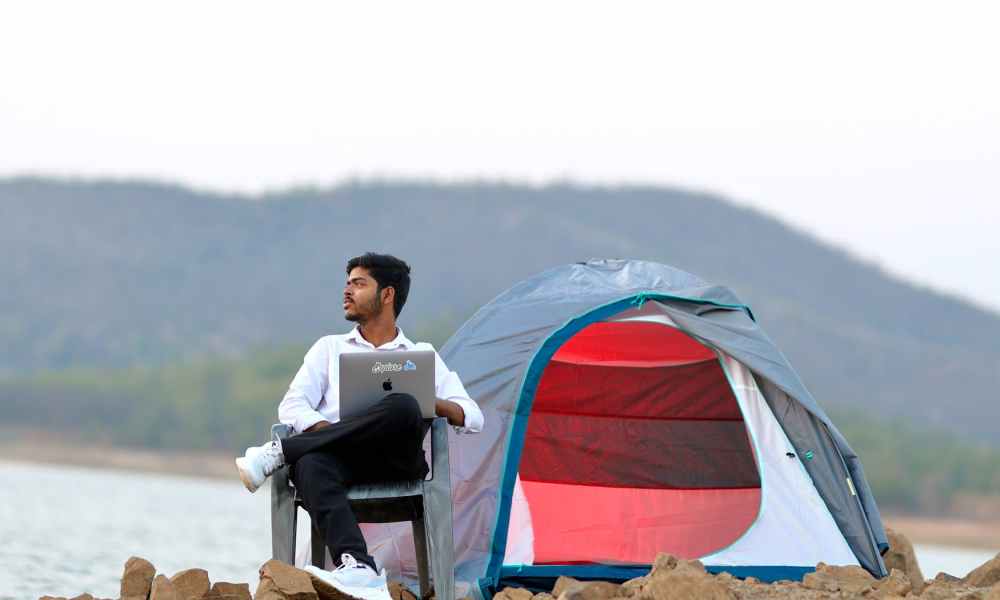 A man sitting while holding a laptop at his campsite off a body of water on a cloudy day