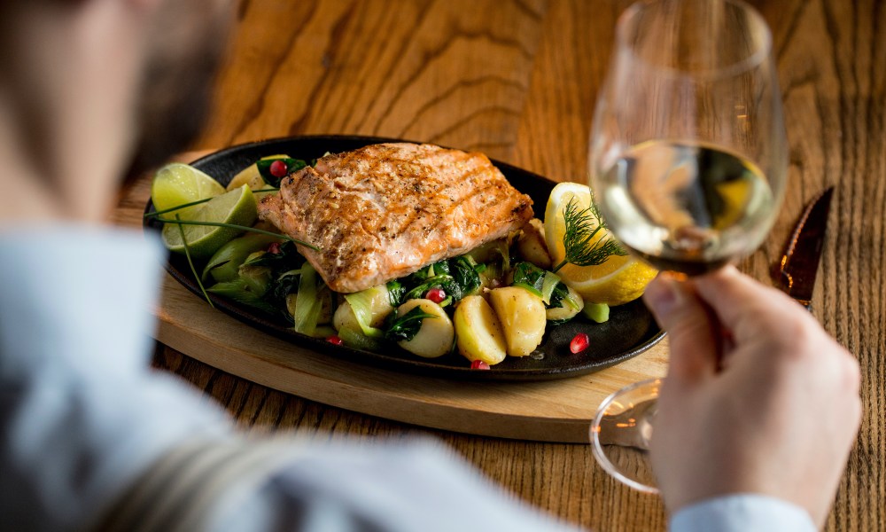 Young man eating salmon fillet with gratinated potatoes, leek and spinach in the restaurant with glass of white wine